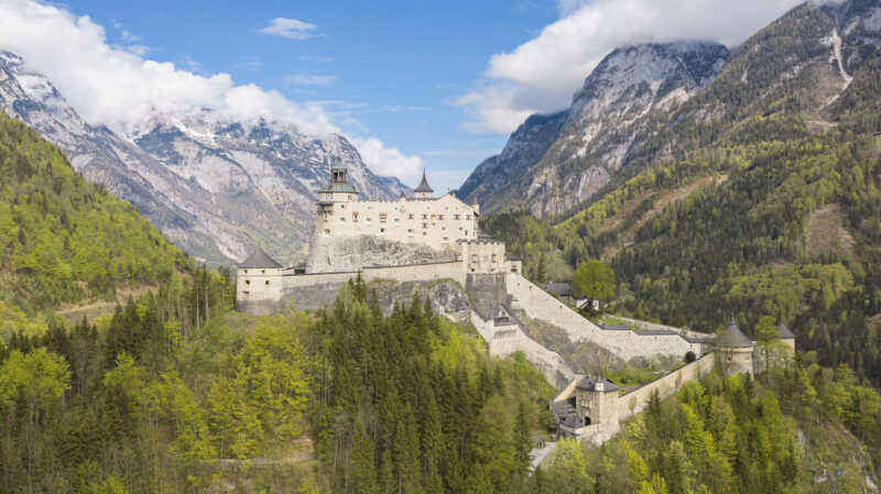 Hohenwerfen Castle perched on a rocky hill, surrounded by lush green forests and mountains under a partly cloudy sky.