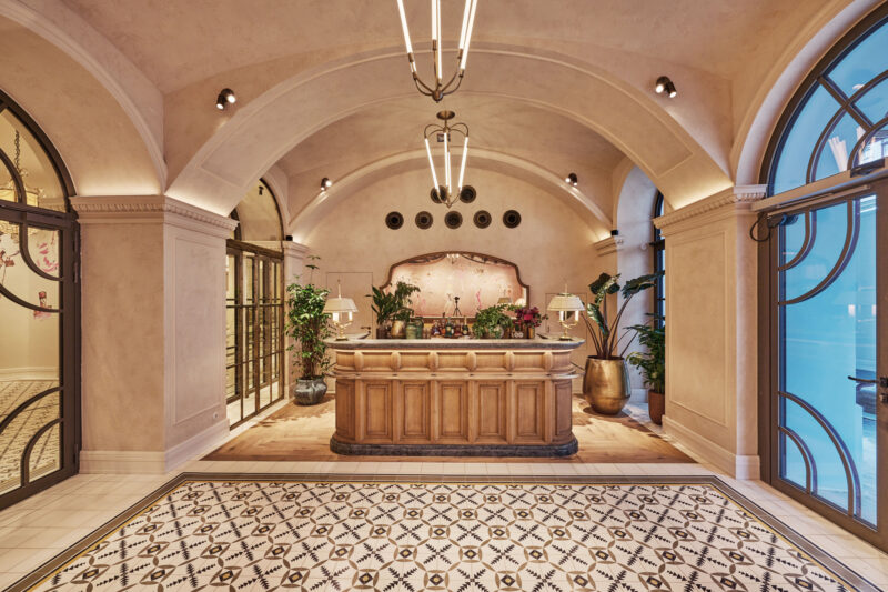 Elegant hotel lobby featuring a wooden reception desk, decorative plants, and ornate tile flooring.