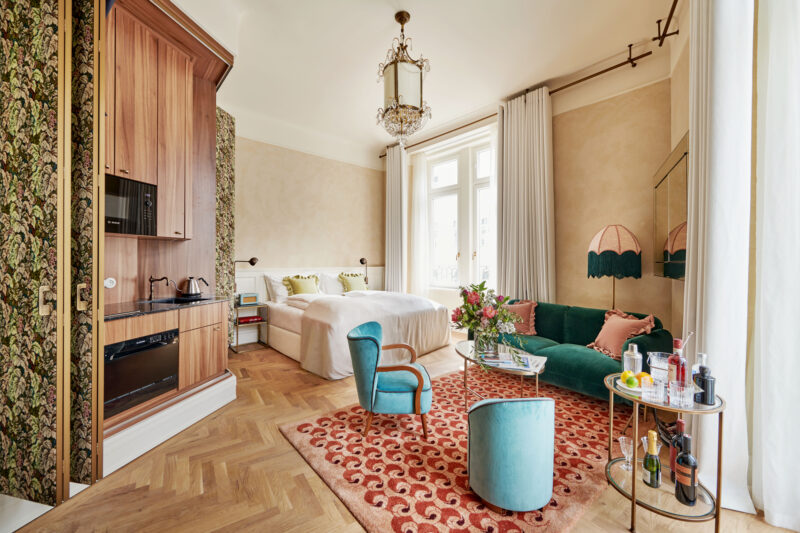 Luxurious hotel suite featuring a green velvet sofa, floral wallpaper, and elegant decor, with a bed and stylish furnishings.