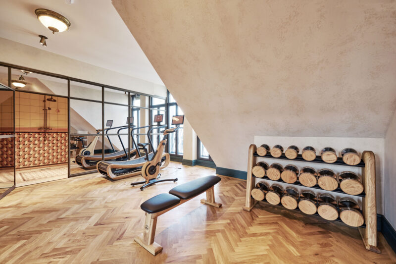 Modern fitness area in Hotel Motto featuring a bench, dumbbell rack, and cardio machines behind glass partitions.