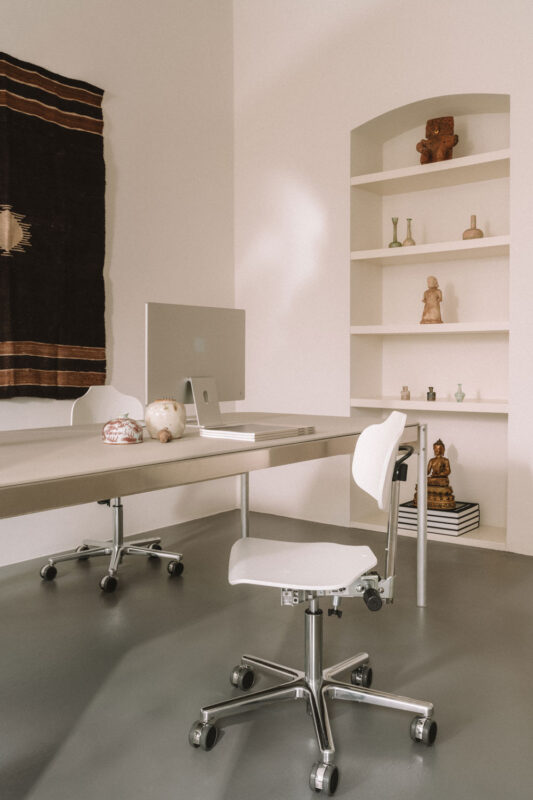 Minimalist office space featuring a desk with a computer, a white chair, and a shelf displaying decorative items.