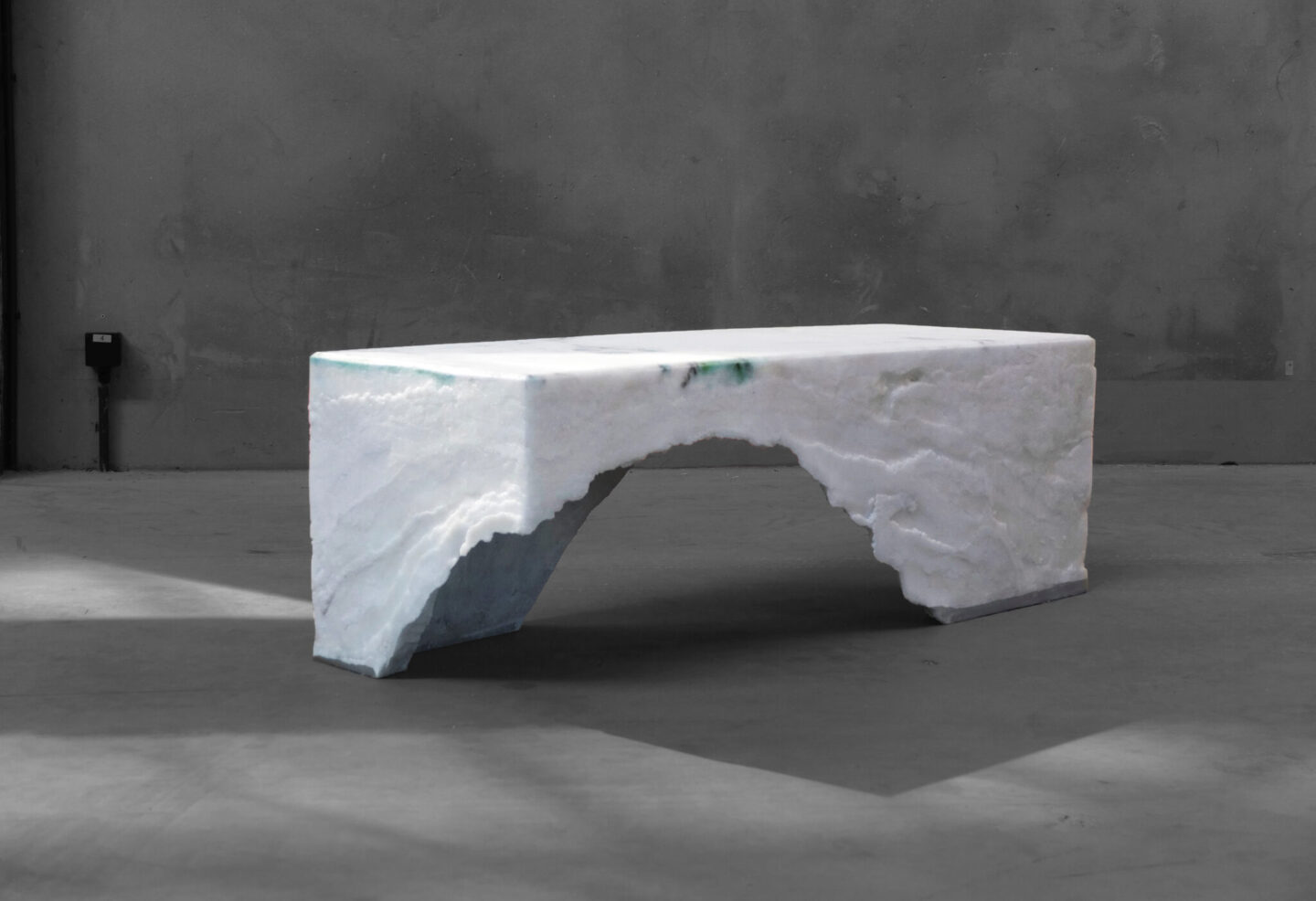 A minimalist white marble bench with an arch design, set against a gray wall and floor.