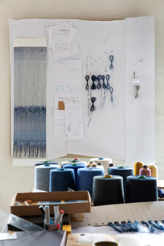 Textured fabric sample in shades of blue, surrounded by sketches and spools of thread on a workspace.