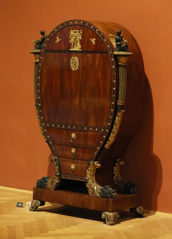 Baroque-style writing cabinet with ornate decorations and lion motifs, set against a red wall.
