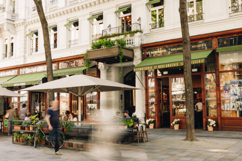 Charming bakery exterior with outdoor seating, people walking by, and greenery in a lively urban setting.