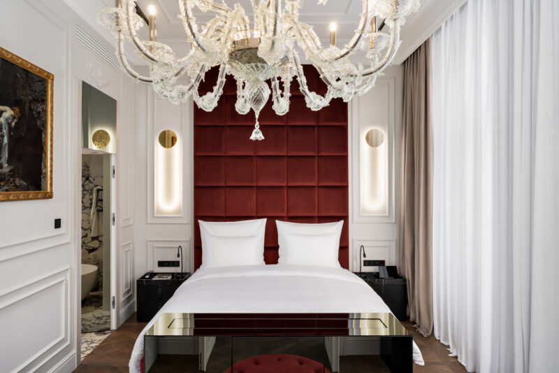 Luxurious hotel room featuring a red tufted headboard, white bedding, a chandelier, and elegant decor.