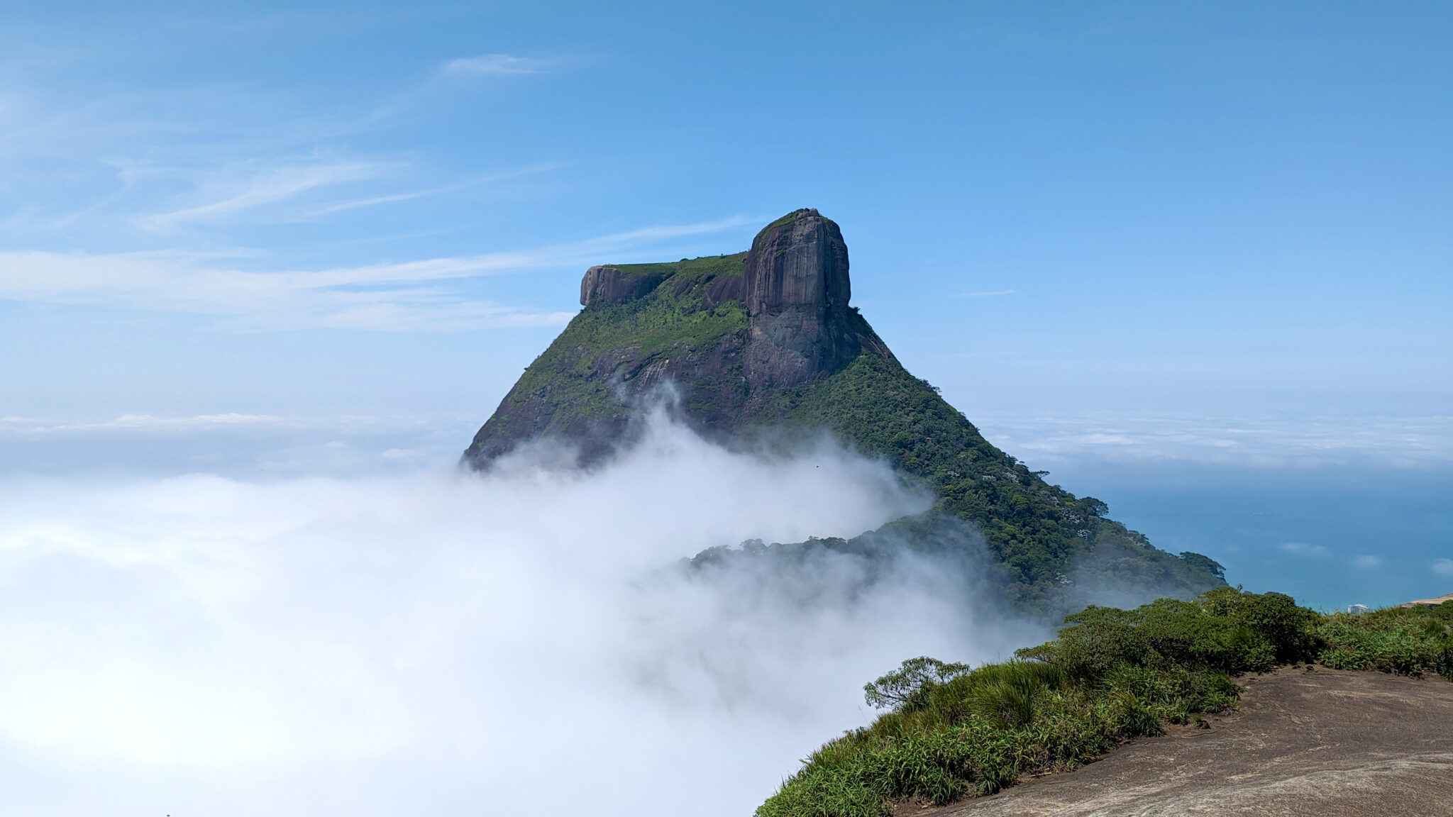 Pedra da Gávea mountain rises above clouds, surrounded by lush greenery and a clear blue sky.