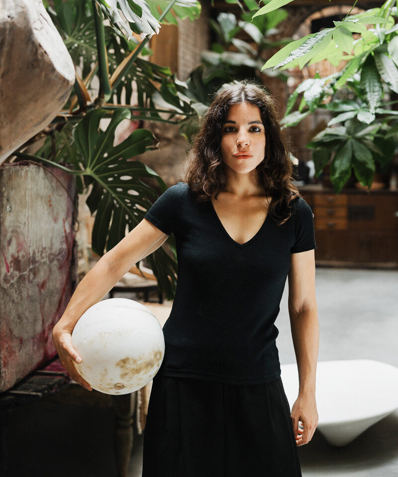 Woman in a black top holding a white ball, surrounded by lush green plants in a stylish indoor setting.