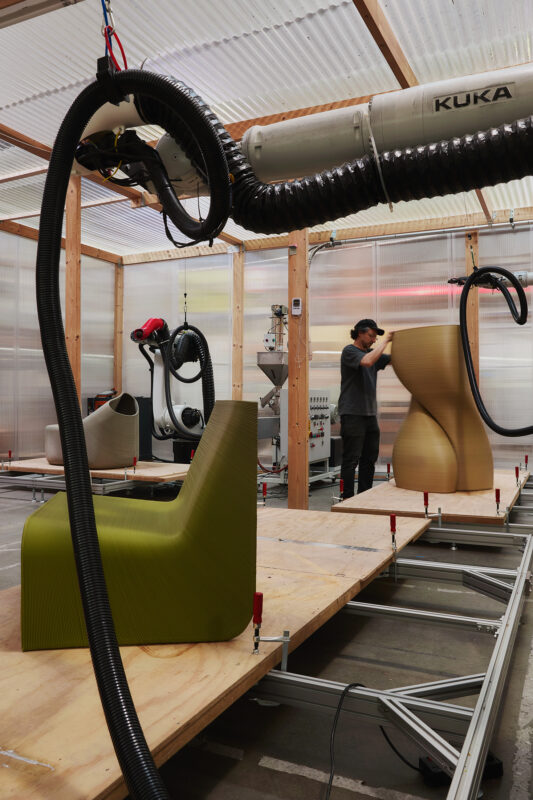 A person operates a robotic arm in a workshop, printing a green chair on a wooden platform.