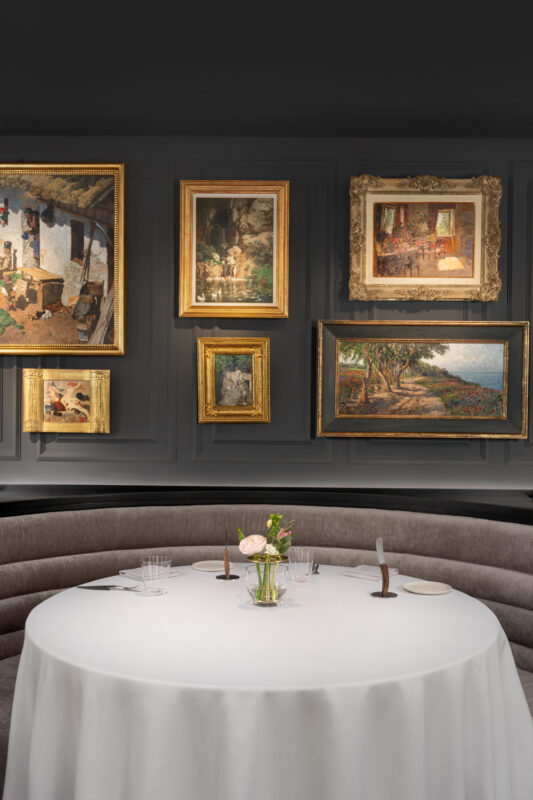 Elegant dining table with a white tablecloth, surrounded by framed artworks on a dark wall in a sophisticated restaurant i...