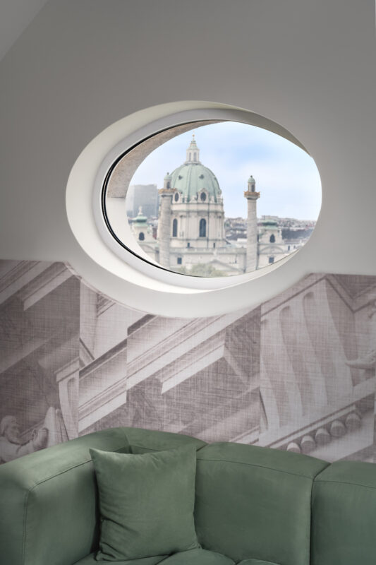 View of a dome and spires through an oval window, with a green sofa and architectural wallpaper in the foreground.