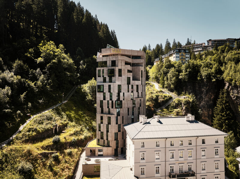 Modern building with a unique facade, surrounded by greenery and mountains, alongside a traditional structure.