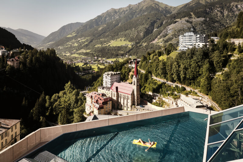 A person relaxes on a float in a rooftop spa pool overlooking the Gasteinertal valley and mountains.