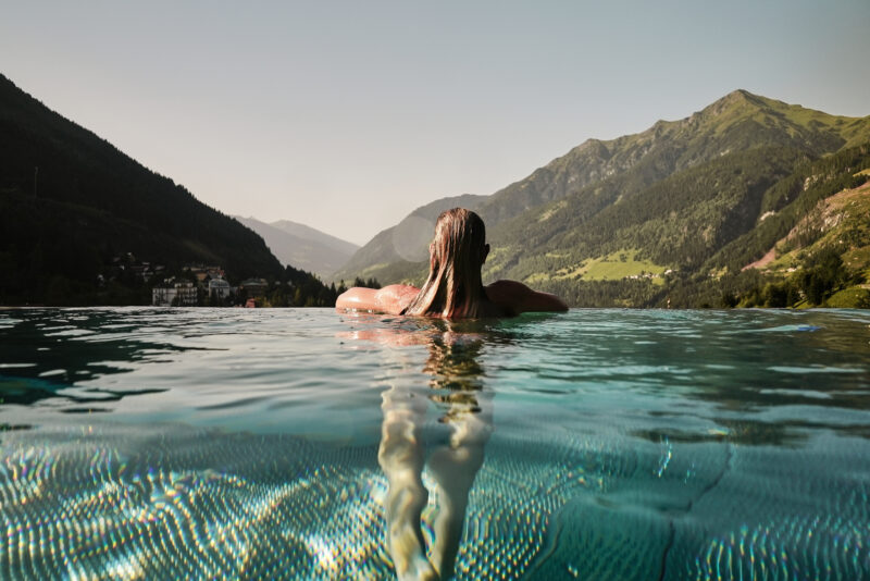 Person relaxing in an infinity pool with mountains and a valley in the background, under a clear sky.