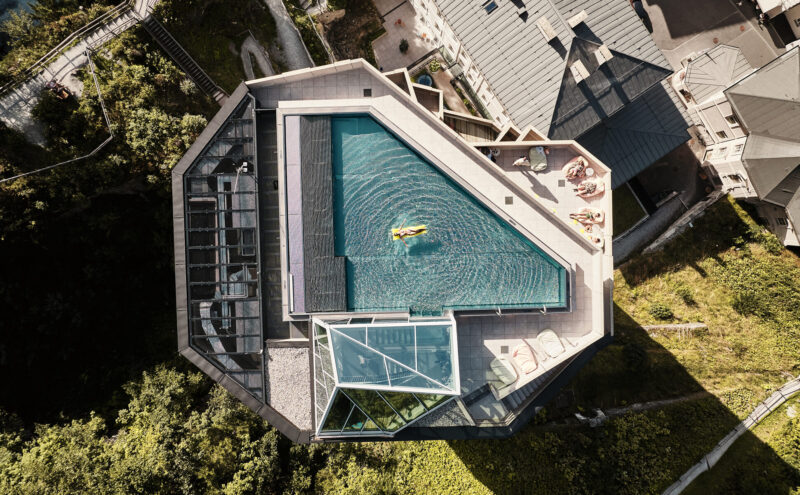 Aerial view of a modern triangular spa pool surrounded by greenery and buildings, with a person floating on the water.