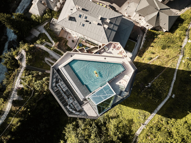 Aerial view of a modern spa pool surrounded by greenery and buildings, with people relaxing by the water.