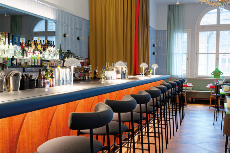 Modern bar interior featuring a long counter, high stools, and a variety of drinks displayed on shelves.