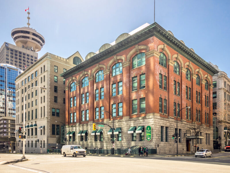 Historic brick building with large windows, adjacent to modern glass structures, under a clear blue sky in Vancouver.