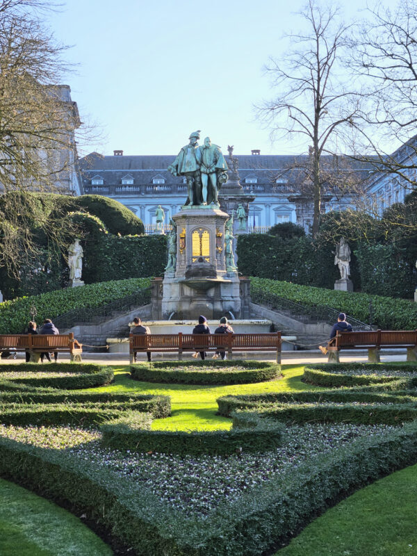 Statue in Petit Sablon Park surrounded by benches and neatly trimmed hedges, with visitors seated in the sun.