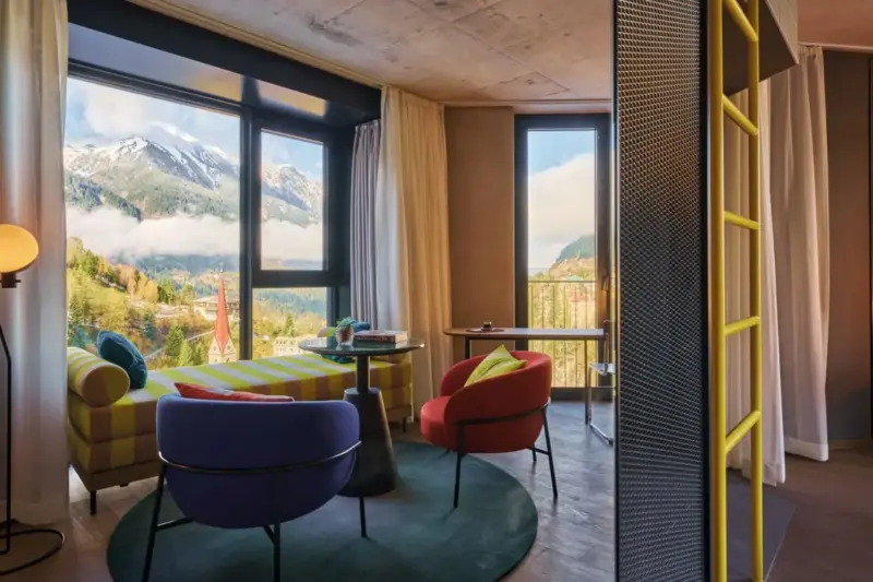 Modern interior of a suite featuring colorful chairs, a panoramic mountain view through large windows, and a cozy atmosphere.