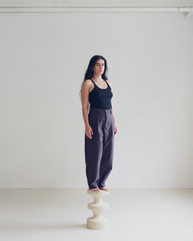 A woman with long dark hair stands barefoot on a sculptural pedestal, wearing a black tank top and loose gray pants.