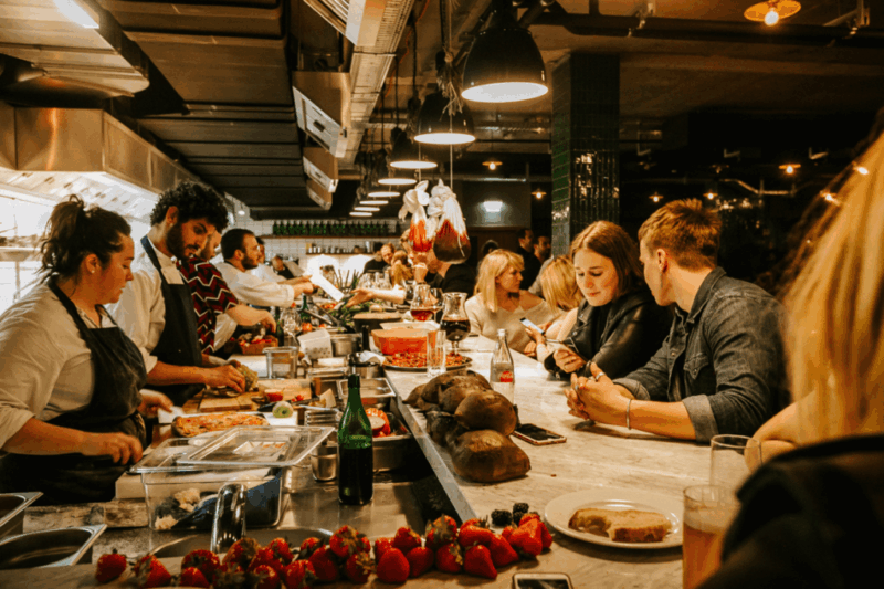 Busy restaurant kitchen scene with chefs preparing food and patrons enjoying drinks at a marble bar.