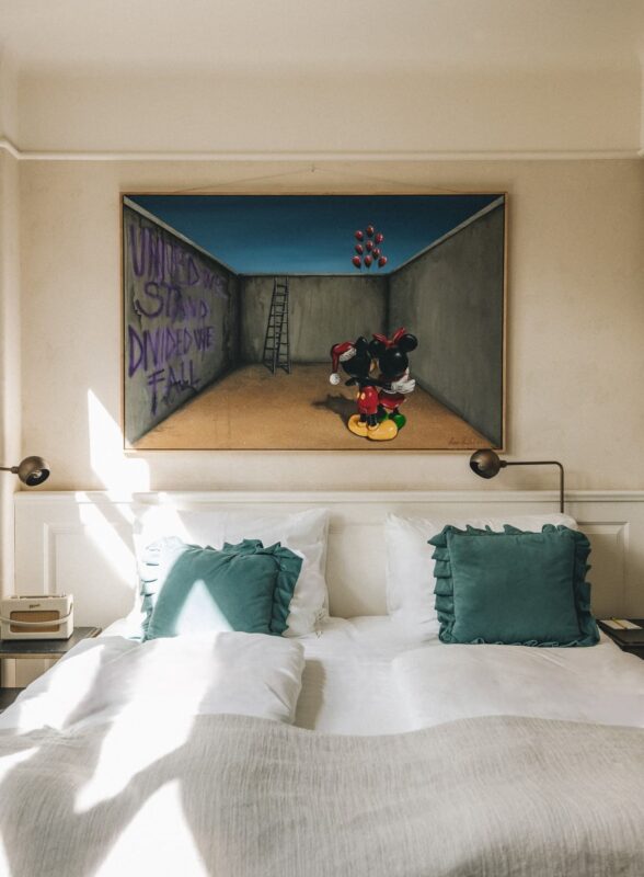 Painting of a cartoon character in a room with graffiti reading "UNITED WE STAND DIVIDED WE FALL" above a bed with teal pi...