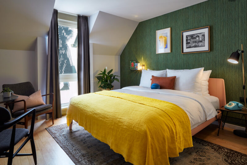Cozy hotel room featuring a bed with a yellow blanket, green wallpaper, and a window with natural light.