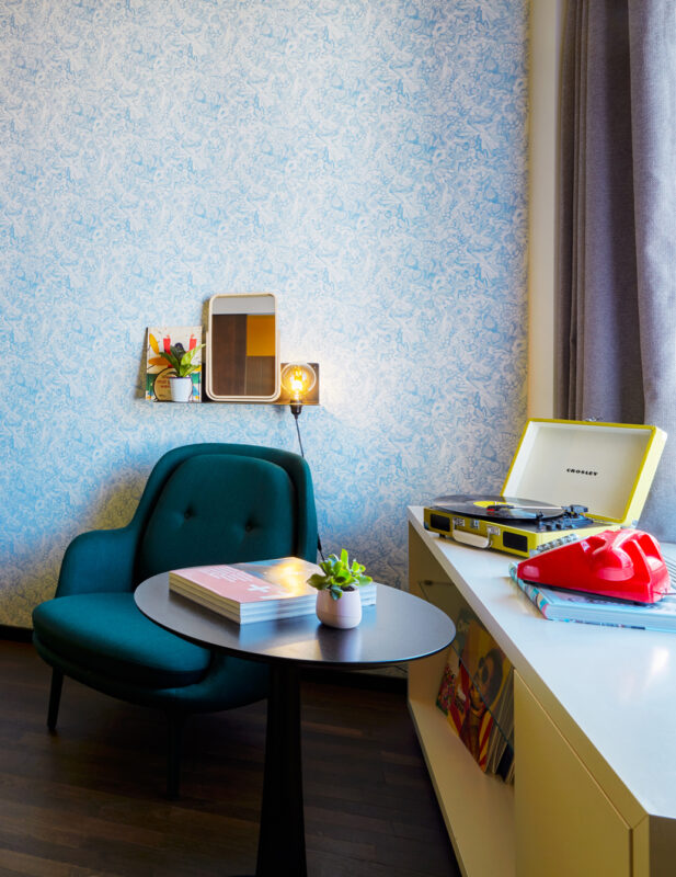 Bright hotel room featuring a yellow bedspread, teal chair, and a record player on a white table against a patterned wall.