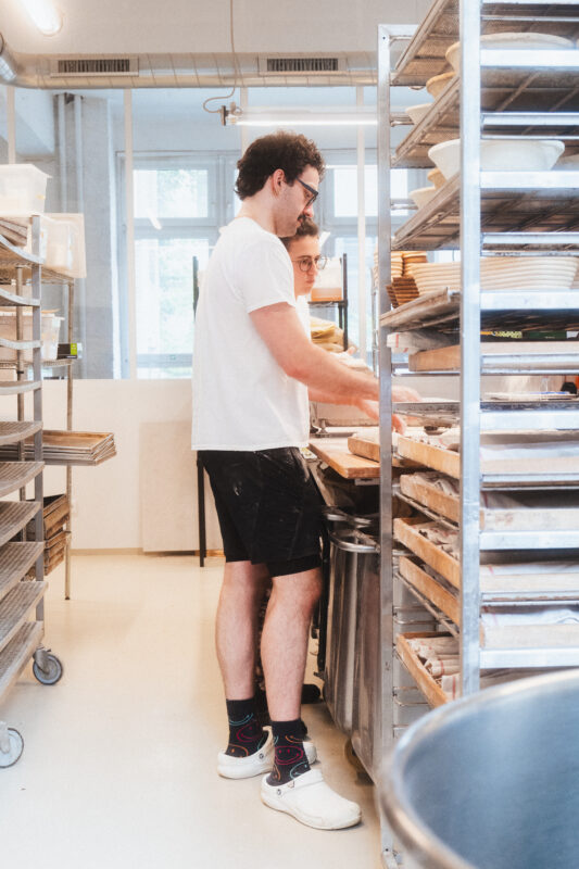 Two bakers working in a bright bakery, one wearing a white shirt and shorts, the other in glasses, surrounded by baking tr...
