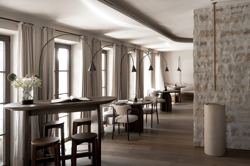 Modern restaurant interior featuring wooden furniture, arched lamps, and large windows with sheer curtains.