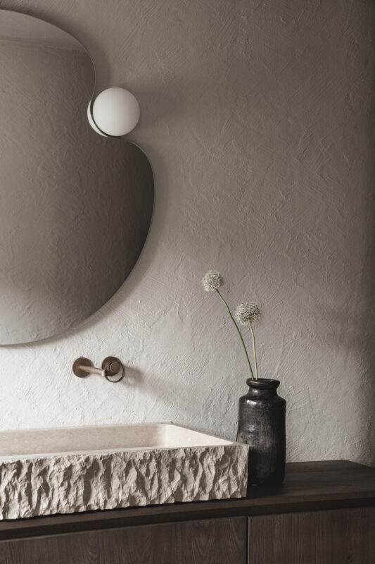 Elegant bathroom interior featuring a textured stone sink, a round mirror, and a dark vase with green flowers.