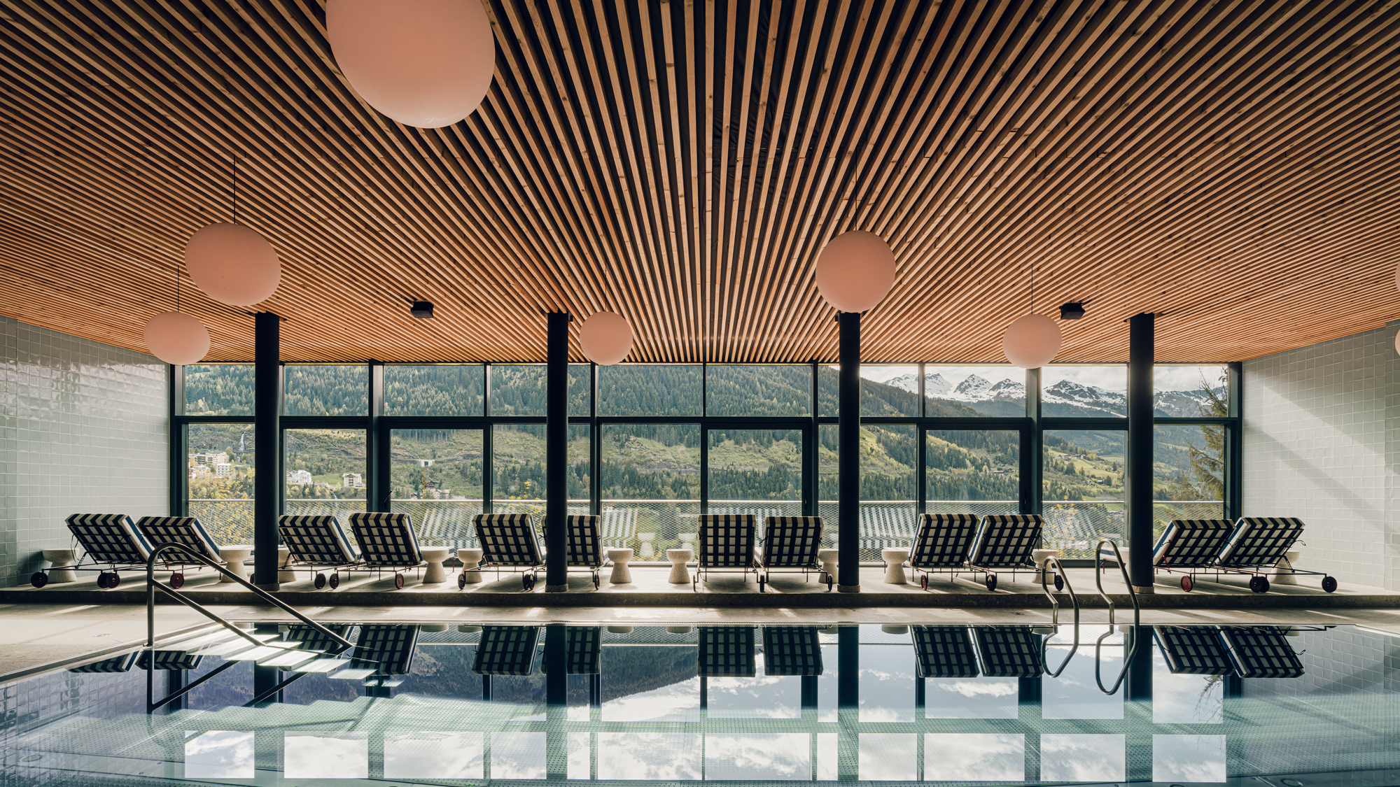 Indoor pool area with lounge chairs, large windows showcasing mountain views, and round pendant lights.
