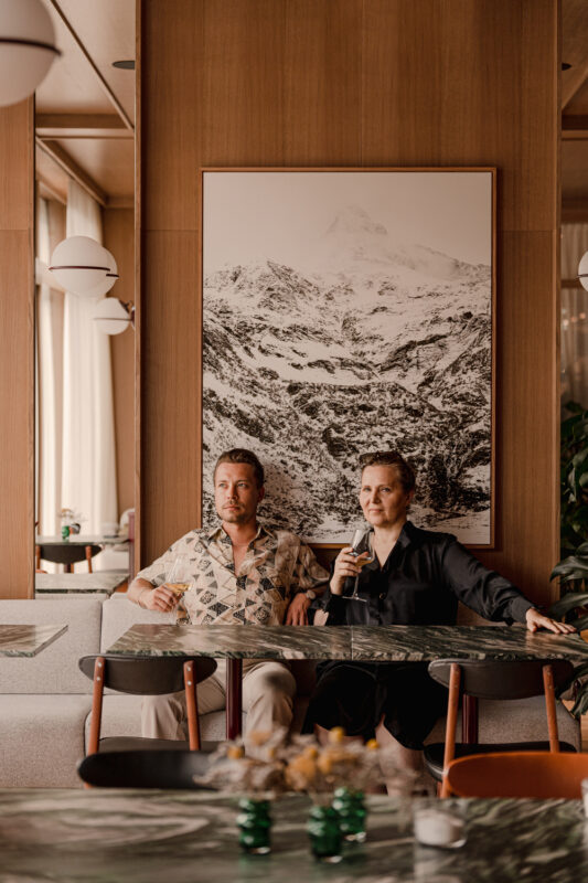 A man and a woman sit at a table in a stylish restaurant, with a mountain landscape photograph on the wall behind them.