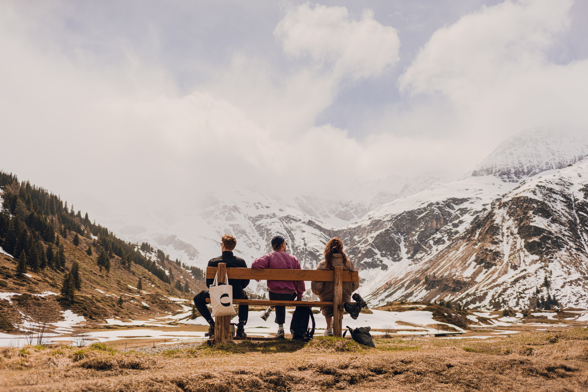 Three people sit on a bench overlooking a snow-capped mountain landscape, with clouds partially obscuring the peaks.
