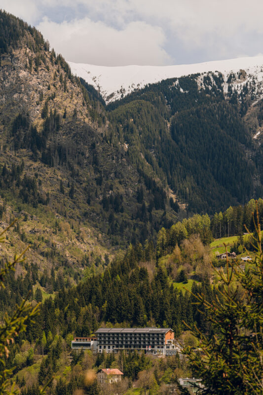 Mountainous landscape featuring a hotel nestled among trees, with snow-capped peaks in the background.