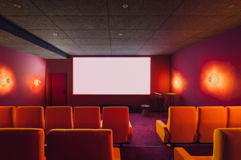 Modern cinema room with orange seating, a large blank screen, and warm lighting on red walls.