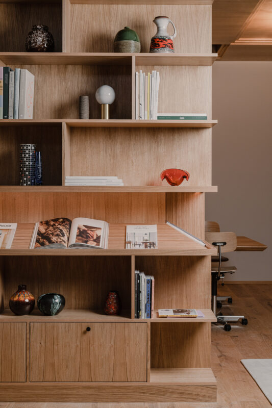 Wooden shelving unit displaying various decorative vases, books, and magazines, with a modern workspace in the background.