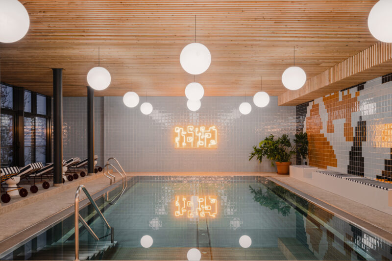 Indoor swimming pool with a wooden ceiling, round lights, and decorative tiles, featuring a neon art piece on the wall.