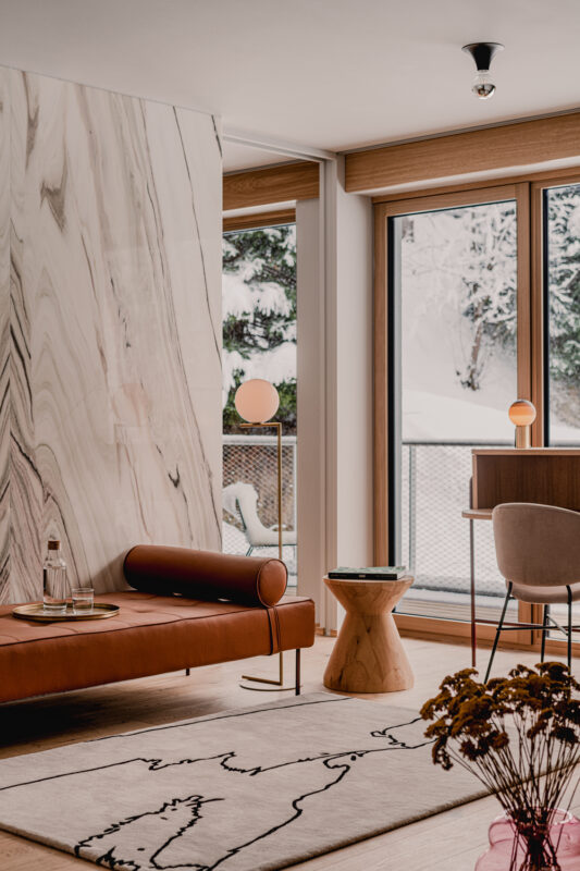 Modern living room featuring a marble wall, a brown leather sofa, a wooden stool, and large windows with a snowy view.