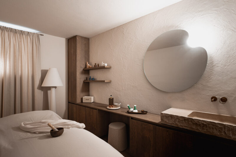 Minimalist interior of a spa-like room featuring a bed, wooden shelves with beauty products, and a round mirror.