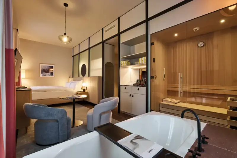 Modern junior suite featuring a freestanding bathtub, sauna, seating area with chairs and table, and a bed in the background.