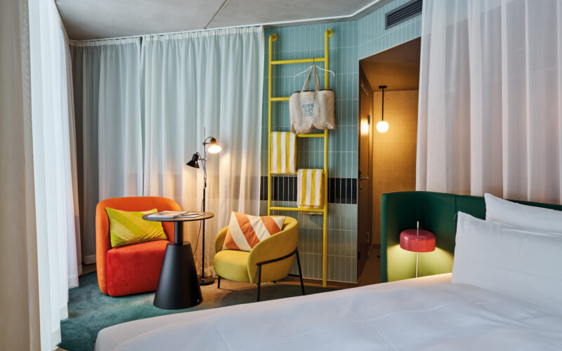 Colorful interior of a hotel double room featuring a bed, chairs, table, and a ladder, with a panoramic view.