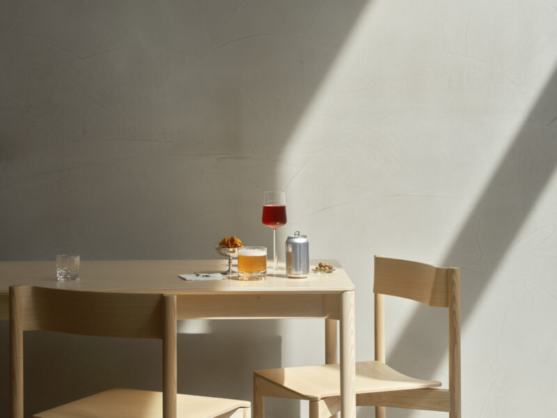 A wooden table with two chairs, featuring a glass of red drink, a cocktail, and a can, illuminated by sunlight.