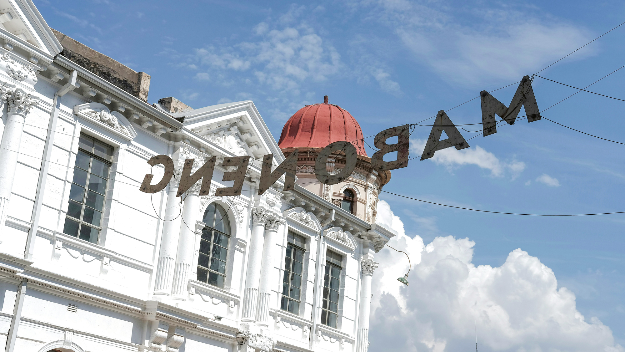 Sign reading "MABONENG" hangs above a white building with ornate architecture and a red dome under a blue sky.