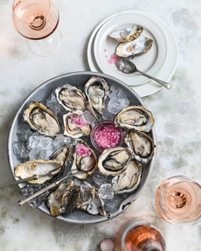 Oysters on ice served with pink pearls in a bowl, accompanied by glasses of rosé wine and a plate.