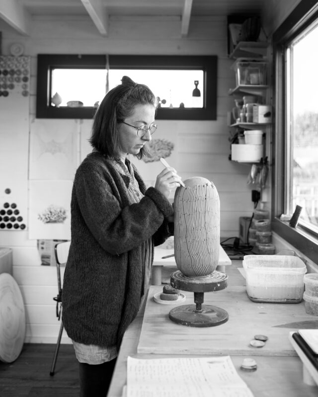 Woman with glasses sculpting a ceramic piece in a bright studio, surrounded by art supplies and natural light.