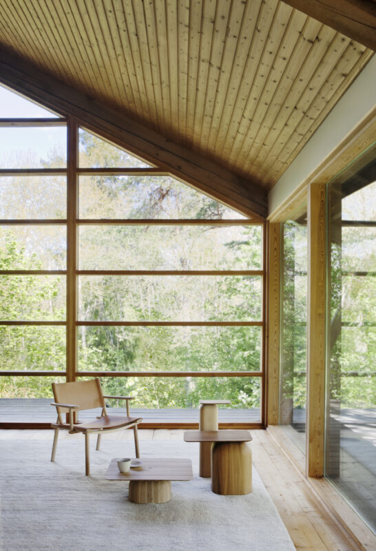 Lounge area featuring a nude ash chair, wooden tables, and large windows with a view of greenery.