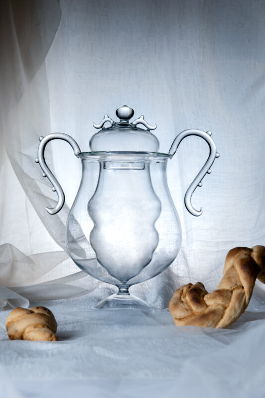 Elegant glass jar with ornate handles, placed on a soft white background alongside twisted bread pieces.