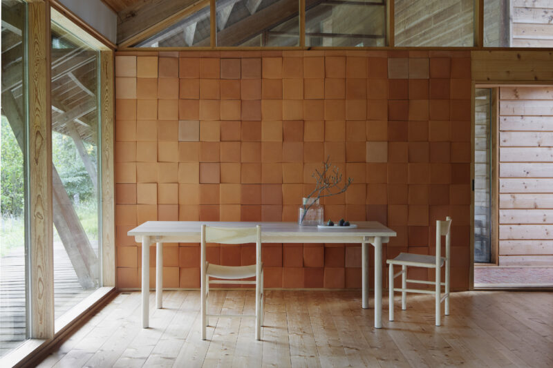 Light wooden table with two Akademia chairs in a room featuring a textured orange wall and large windows.